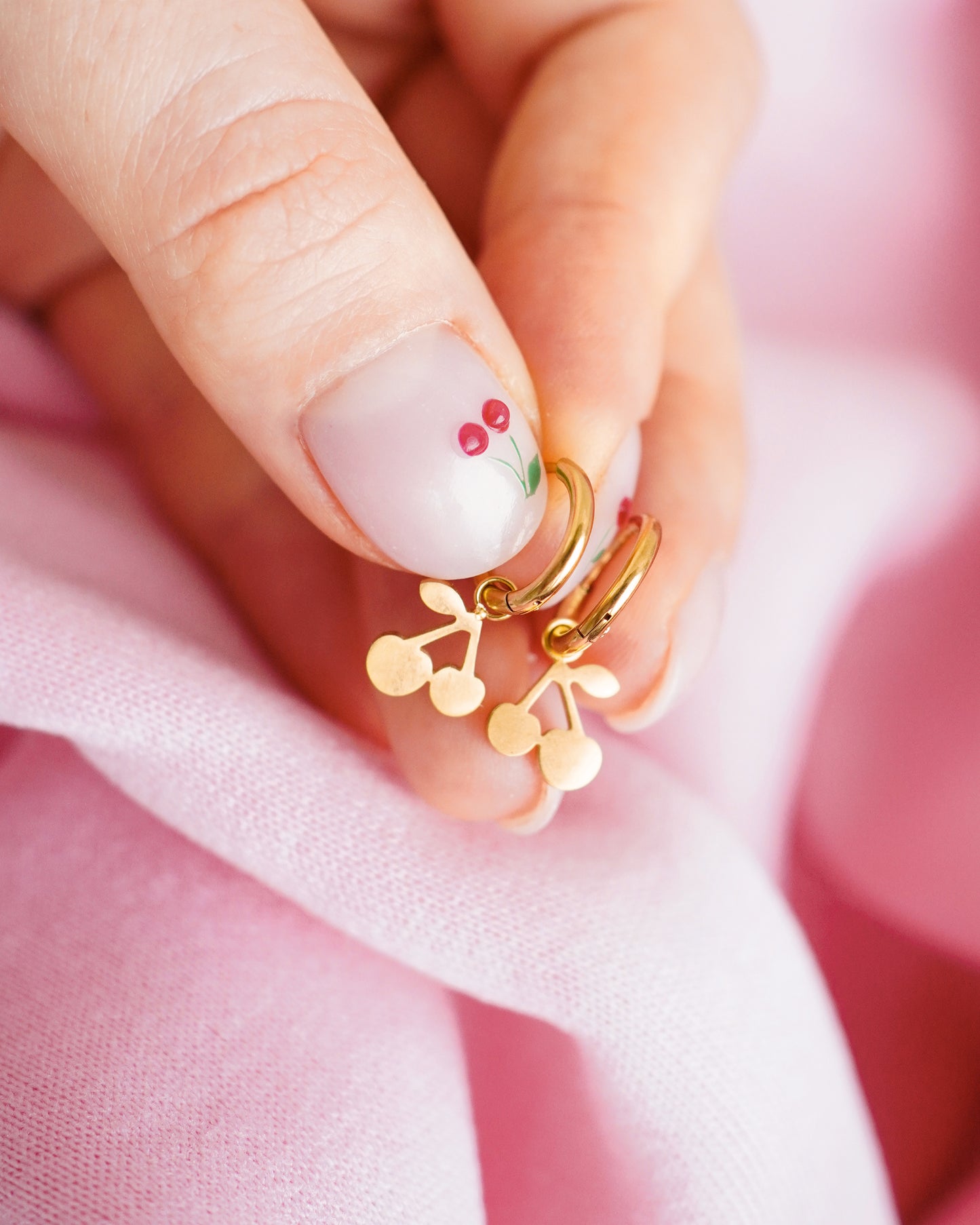 Gold earrings with cherry design held between fingers against a pink fabric background