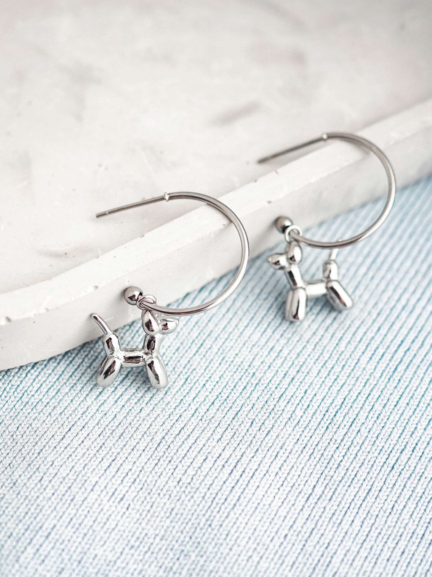 Silver stud earrings with balloon dog charms on a light blue fabric background