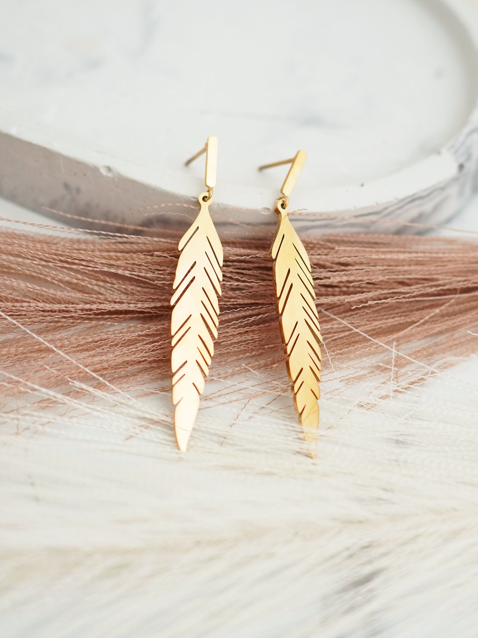 Gold-plated  feather-shaped, stud earrings on a marble surface