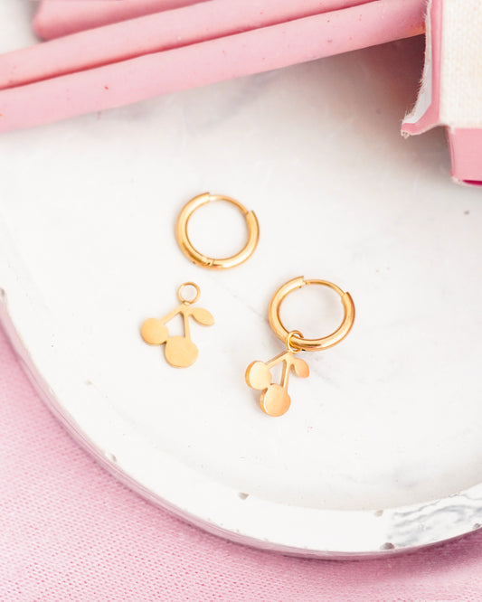 Gold hoop earrings with cherry charms on a white plate with pink background