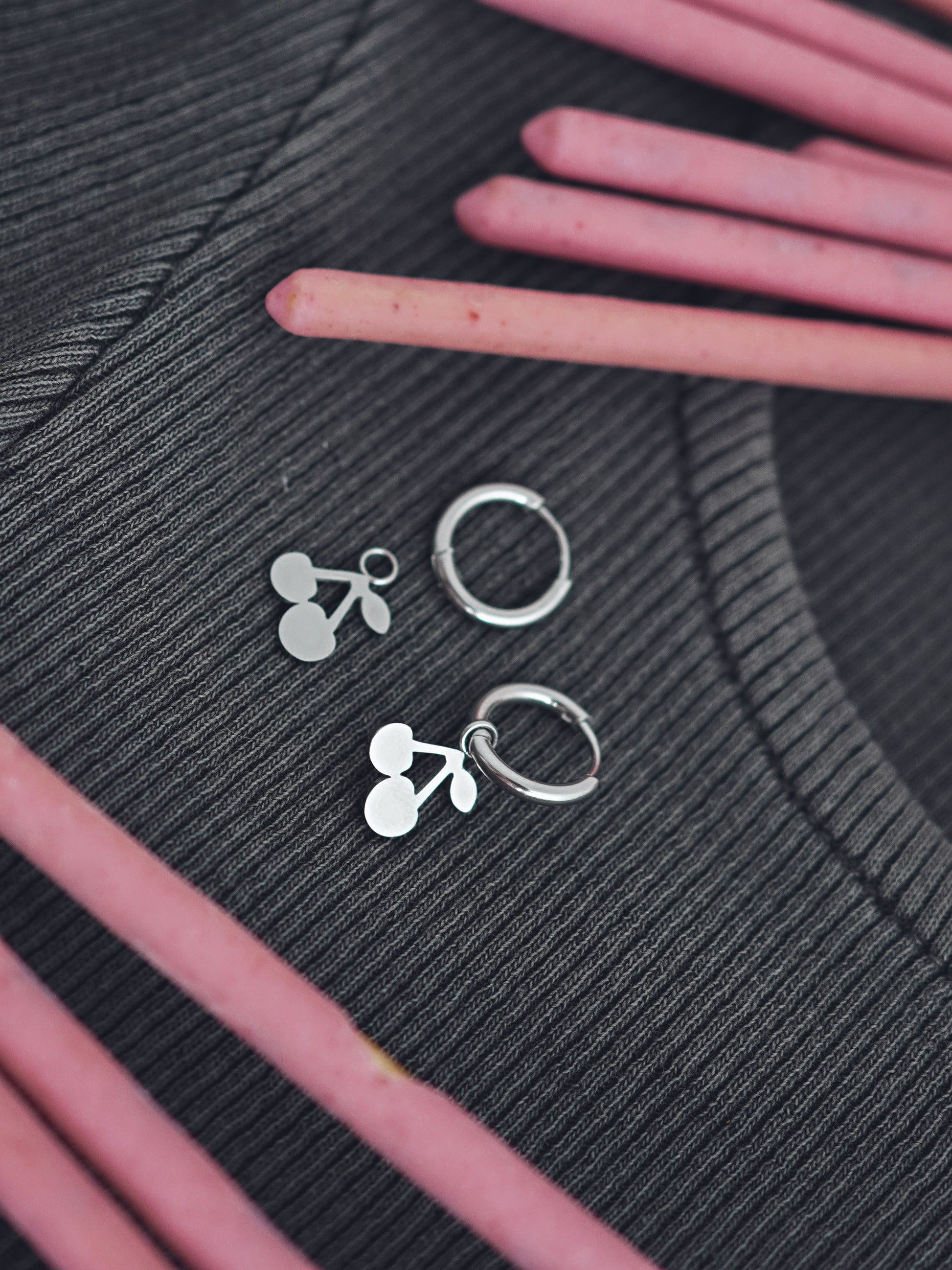 Silver colored cherry-shaped earrings on a textured black surface with pink sticks.