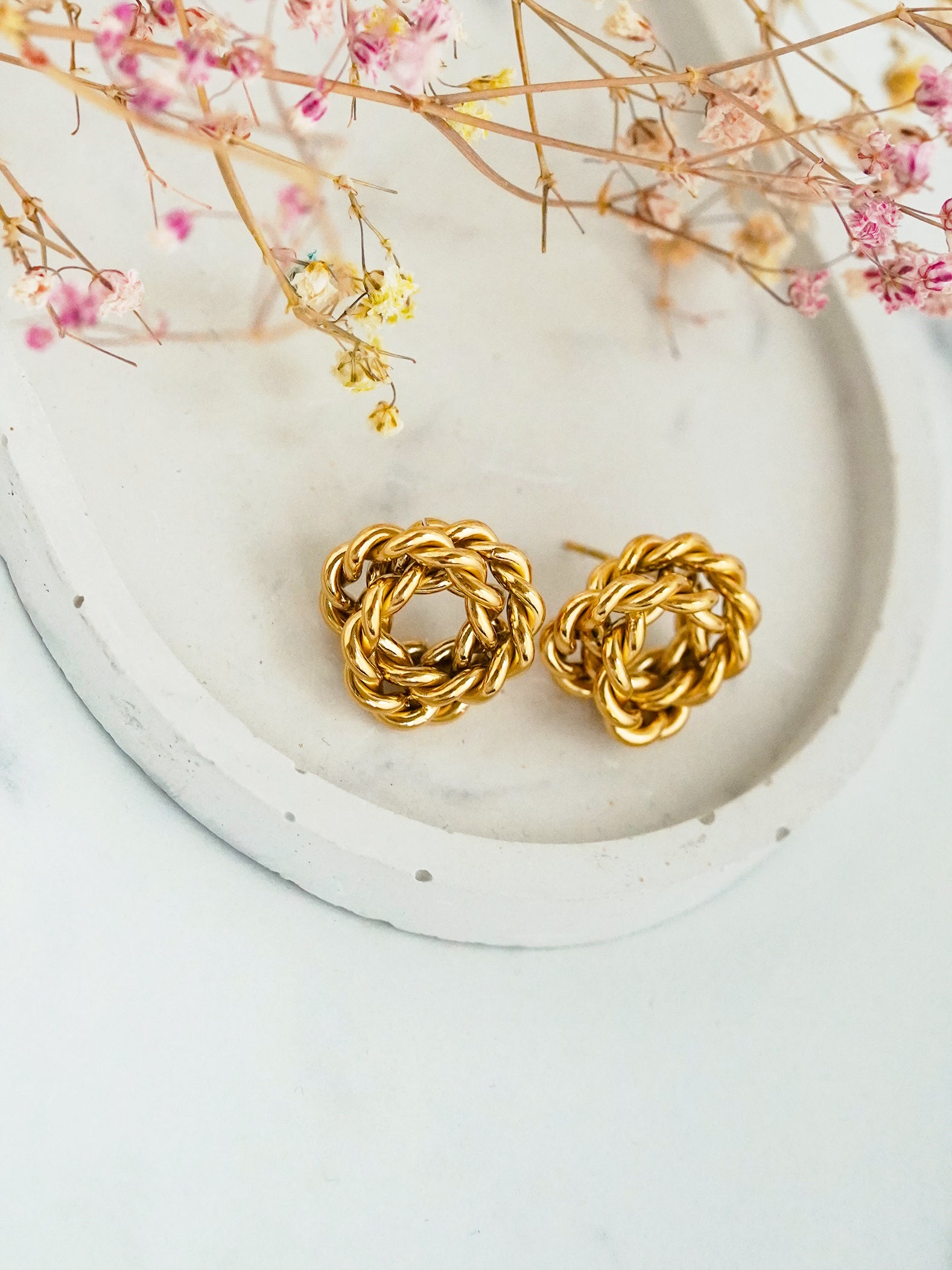 Gold-plated earrings on a white plate with dried flowers in the background