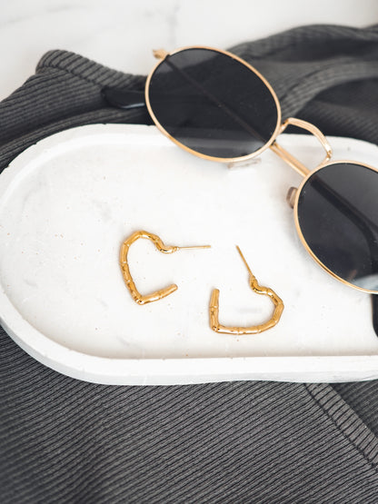 Gold colored heart-shaped earrings on a white surface with sunglasses in the background