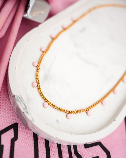 Gold colored necklace with pink beads on a white stand against a pink background