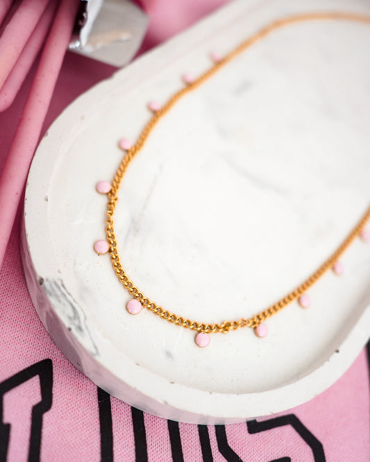 Gold colored necklace with pink beads on a white stand against a pink background