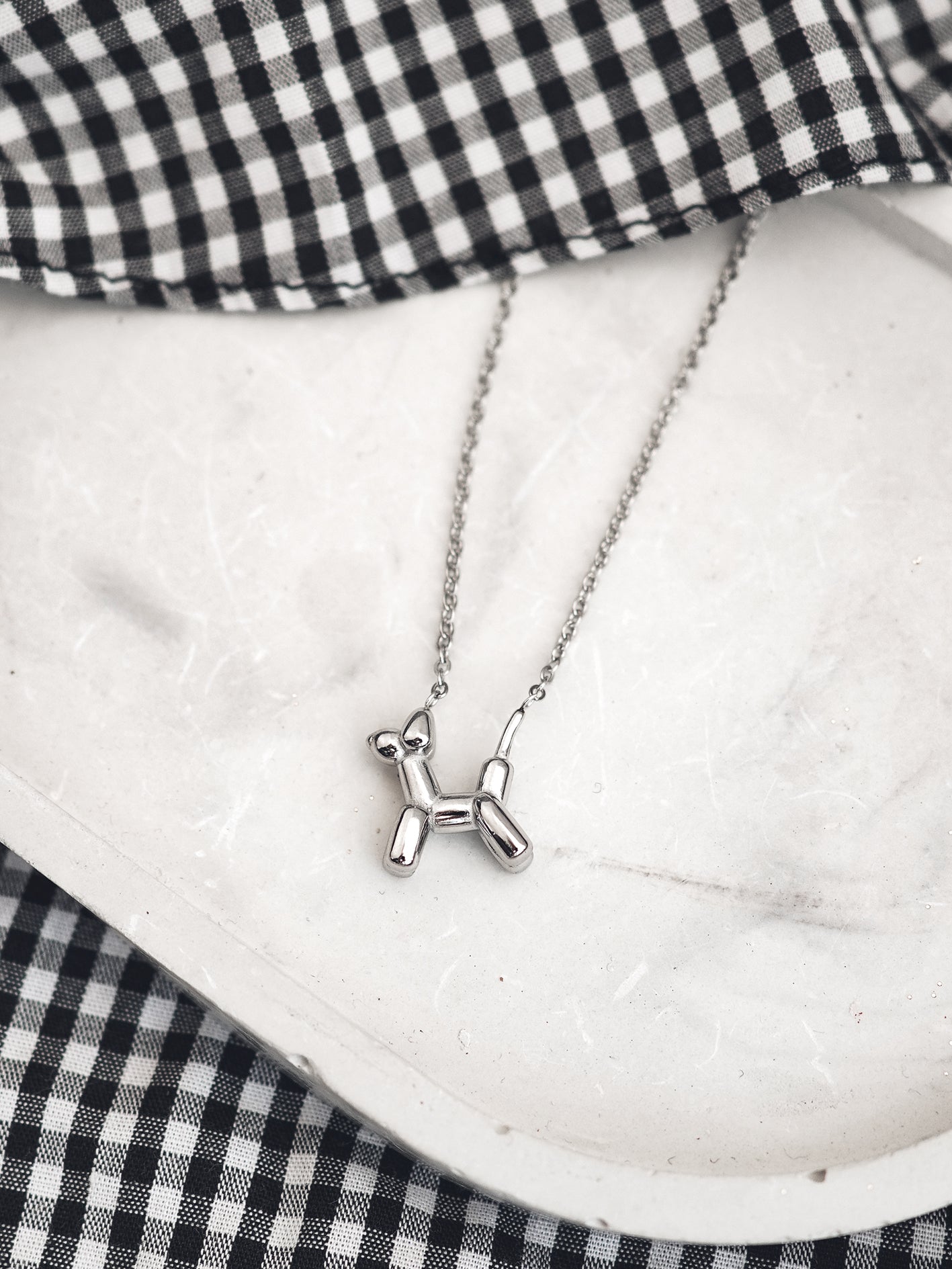 Silver balloon dog necklace on a white surface with a black and white checkered fabric background